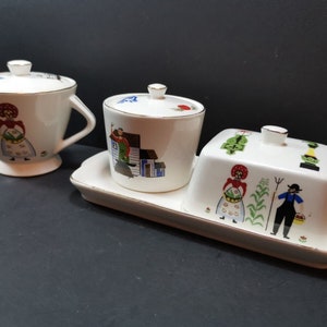 May include: A white ceramic butter dish with a lid and two matching sugar bowls with lids. The dishes are decorated with a folk art design featuring a woman in a red dress, a man in blue overalls, and a house.