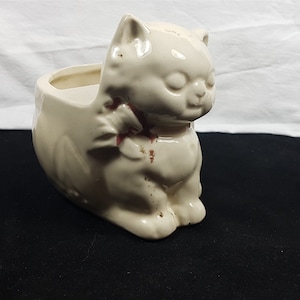 May include: A white ceramic cat planter with a brown painted detail on the chest. The planter is shaped like a sitting cat with a pot for plants in the back.