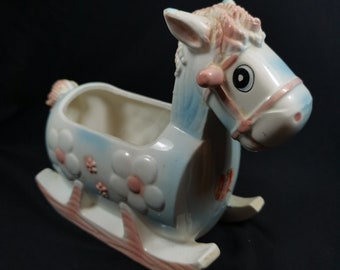 May include: A blue and white ceramic rocking horse planter with pink flowers and a pink bridle. The horse has a white face with a black eye and a pink nose.