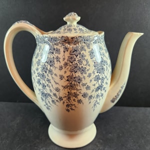 May include: Cream-colored ceramic teapot with a blue floral pattern. The teapot has a curved handle, a spout, and a lid with a small knob. The floral design covers the body of the teapot, lid, and spout. The teapot sits on a small, round base.