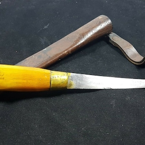 May include: A knife with a wooden handle and a curved blade, accompanied by a brown leather sheath. The handle is light yellow with a brass collar and a small emblem. The sheath has a leather strap.