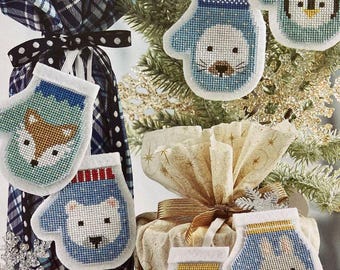 Peut inclure: Ornements de Noël en forme de mitaines, chacun avec un motif animalier différent. Les ornements sont bleus, blancs et marron, avec un aspect cousu. Certains sont accrochés à un sapin de Noël, d'autres près de cadeaux emballés.