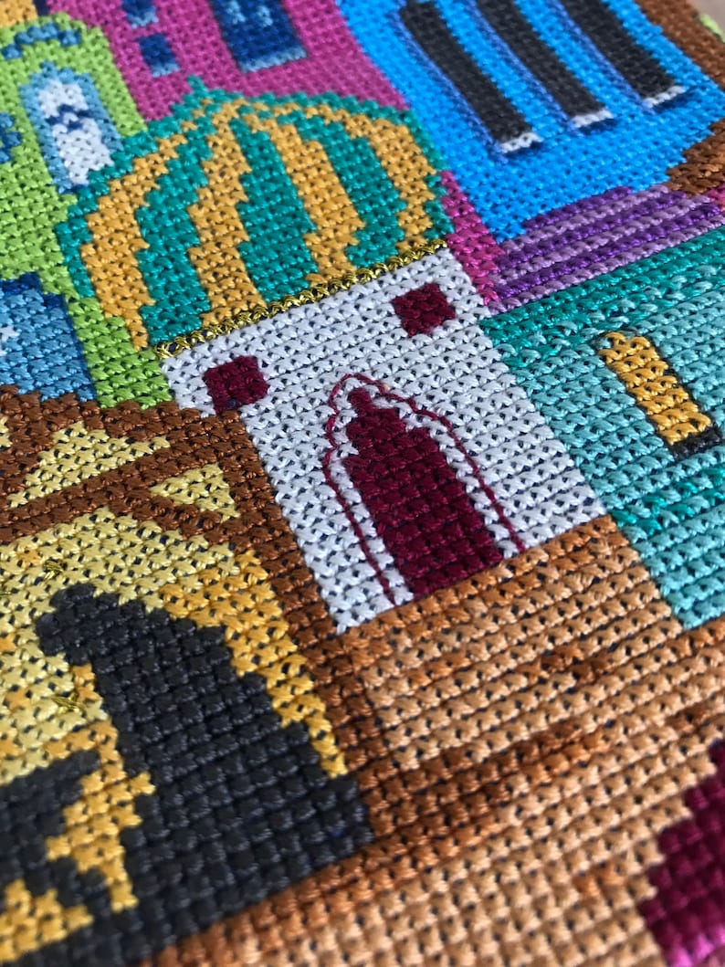 Peut inclure: Gros plan sur une broderie au point de croix color&eacute;e repr&eacute;sentant une ville avec des b&acirc;timents, un d&ocirc;me et une vari&eacute;t&eacute; de couleurs, notamment le vert, le jaune, le bleu, le violet et le marron.