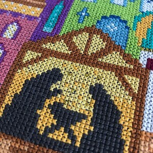 Peut inclure: Gros plan sur une broderie au point de croix repr&eacute;sentant un b&acirc;timent brun avec un toit jaune et une silhouette noire d'une personne &agrave; l'int&eacute;rieur. Le b&acirc;timent est entour&eacute; d'autres b&acirc;timents et motifs color&eacute;s.