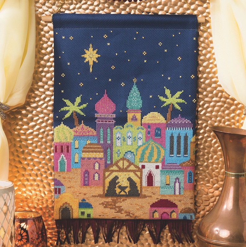 Peut inclure: Une tapisserie murale au point de croix repr&eacute;sentant une sc&egrave;ne nocturne d'une ville avec une &eacute;toile au-dessus. La ville est compos&eacute;e de b&acirc;timents color&eacute;s avec une cr&egrave;che au centre. L'arri&egrave;re-plan est bleu fonc&eacute; avec des &eacute;toiles dor&eacute;es.