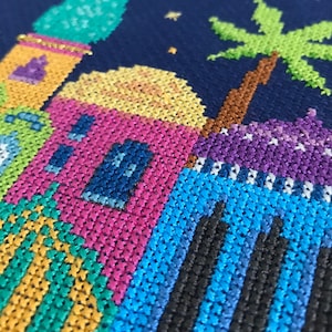 Peut inclure: Une broderie au point de croix color&eacute;e repr&eacute;sentant une sc&egrave;ne nocturne avec un palmier et une rang&eacute;e de maisons aux d&eacute;tails bleus, verts, jaunes, roses et violets.
