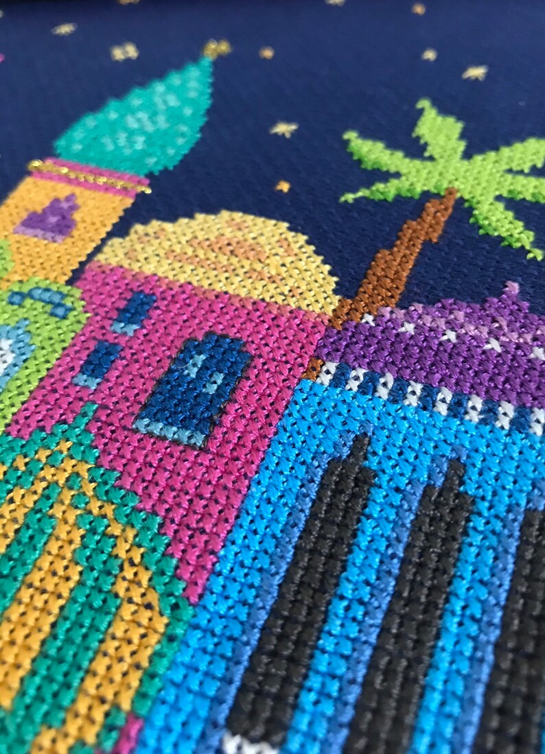 Peut inclure: Une broderie au point de croix color&eacute;e repr&eacute;sentant une sc&egrave;ne nocturne avec un palmier et une rang&eacute;e de maisons aux d&eacute;tails bleus, verts, jaunes, roses et violets.