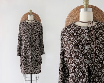May include: A long-sleeved, brown dress with a floral pattern. The dress features a round neckline, decorative buttons, and a tie closure. The dress is displayed on a mannequin with a woven hat.