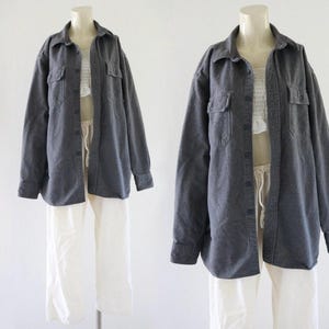 以下が含まれることがあります： 2体のマネキンがカジュアルな服装を展示しています。各マネキンは、2つの胸ポケットが付いた濃い灰色のボタンダウンシャツと白いパンツを着用しています。シャツは開いており、白いトップスが見えています。この服装はシンプルでリラックスしています。