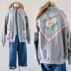 May include: A gray sweatshirt jacket with floral and gingham pattern accents. The jacket features a light blue trim, pockets, and a floral design on the back. The outfit includes blue jeans and a straw hat.