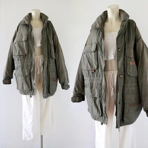 Pu&ograve; includere: Due giacche verde oliva con tasche multiple e vita con coulisse sono esposte su manichini. Le giacche hanno un colletto alto e chiusure con bottoni. I manichini indossano top e pantaloni bianchi.