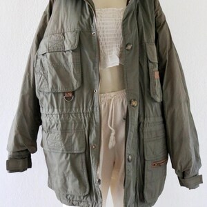 Pu&ograve; includere: Giacca utility verde oliva con pi&ugrave; tasche, chiusura con zip e bottoni e vita con coulisse. La giacca ha un collo alto ed &egrave; esposta su un manichino. La giacca &egrave; in stile vintage.