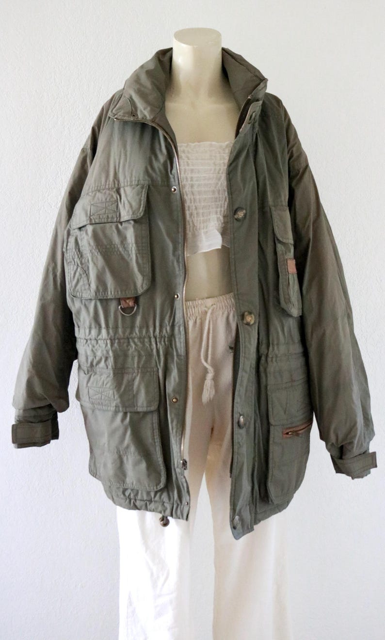 Pu&ograve; includere: Giacca utility verde oliva con pi&ugrave; tasche, chiusura con zip e bottoni e vita con coulisse. La giacca ha un collo alto ed &egrave; esposta su un manichino. La giacca &egrave; in stile vintage.