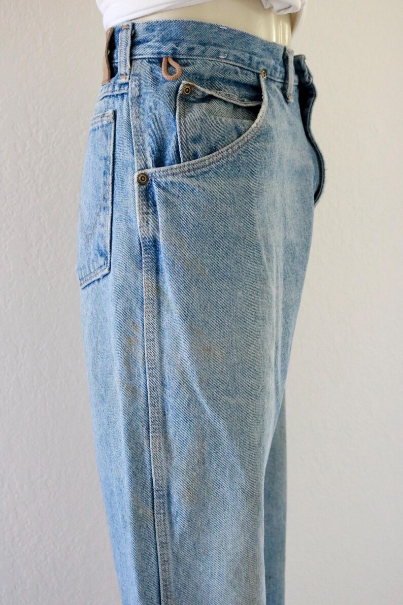 May include: Light blue denim jeans with a classic five-pocket design. The jeans have a high waist and a straight leg cut. Features include metal rivets and a brown leather belt loop. The denim has a faded wash with subtle wear marks.