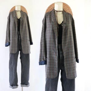 May include: A plaid blazer in shades of gray, brown, and blue, featuring a single button closure and cuffed sleeves. The blazer is layered over a black top and paired with dark gray denim jeans, displayed on a mannequin with a straw hat.