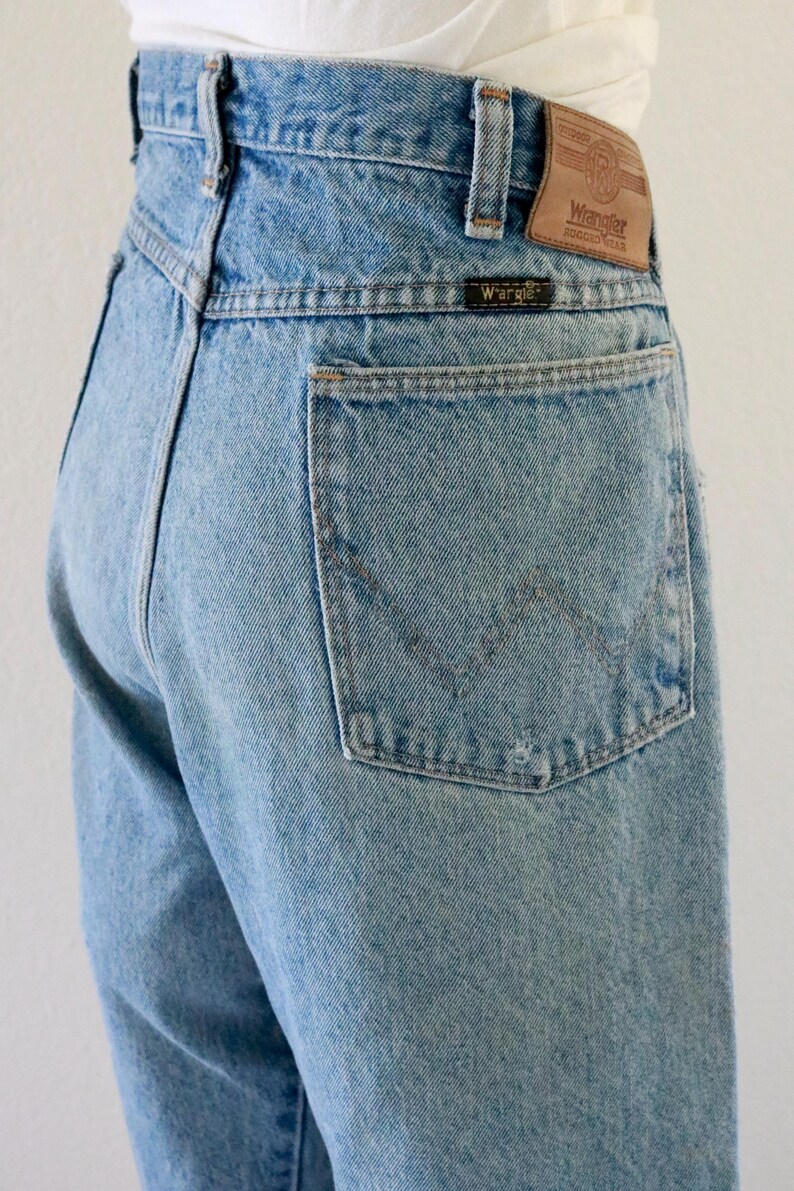 May include: Light blue denim jeans with a classic five-pocket design. The jeans feature a Wrangler brand patch on the back waistband and a small Wrangler label on the back pocket. The jeans are a light wash and have a straight leg.