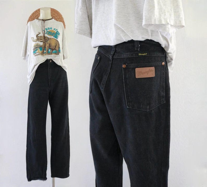 Puede incluir: Vaqueros Wrangler negros con un parche de cuero marr&oacute;n en el bolsillo trasero. Los vaqueros se exhiben en un maniqu&iacute; con una camiseta blanca con un gr&aacute;fico de elefante. La camiseta tiene el texto "Bug At".