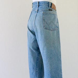 May include: Light blue denim jeans with a high waist and a straight leg. A straw hat with a brown band sits atop a white top. The jeans have a classic five-pocket design and a leather patch.