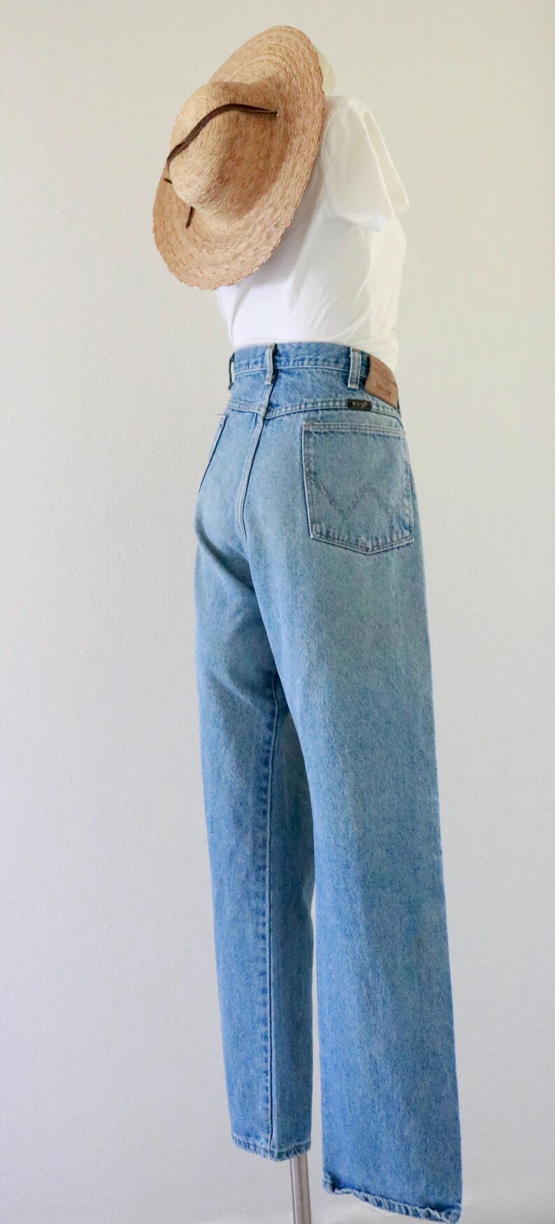 May include: Light blue denim jeans with a high waist and a straight leg. A straw hat with a brown band sits atop a white top. The jeans have a classic five-pocket design and a leather patch.