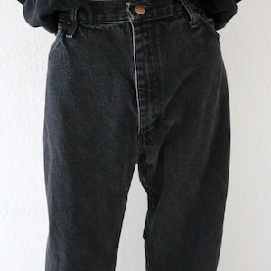 May include: Black denim jeans with a button closure and a straight leg fit. The jeans are worn with a black t-shirt that has the text "The Maya Meetings at Texas 1994" printed on it.