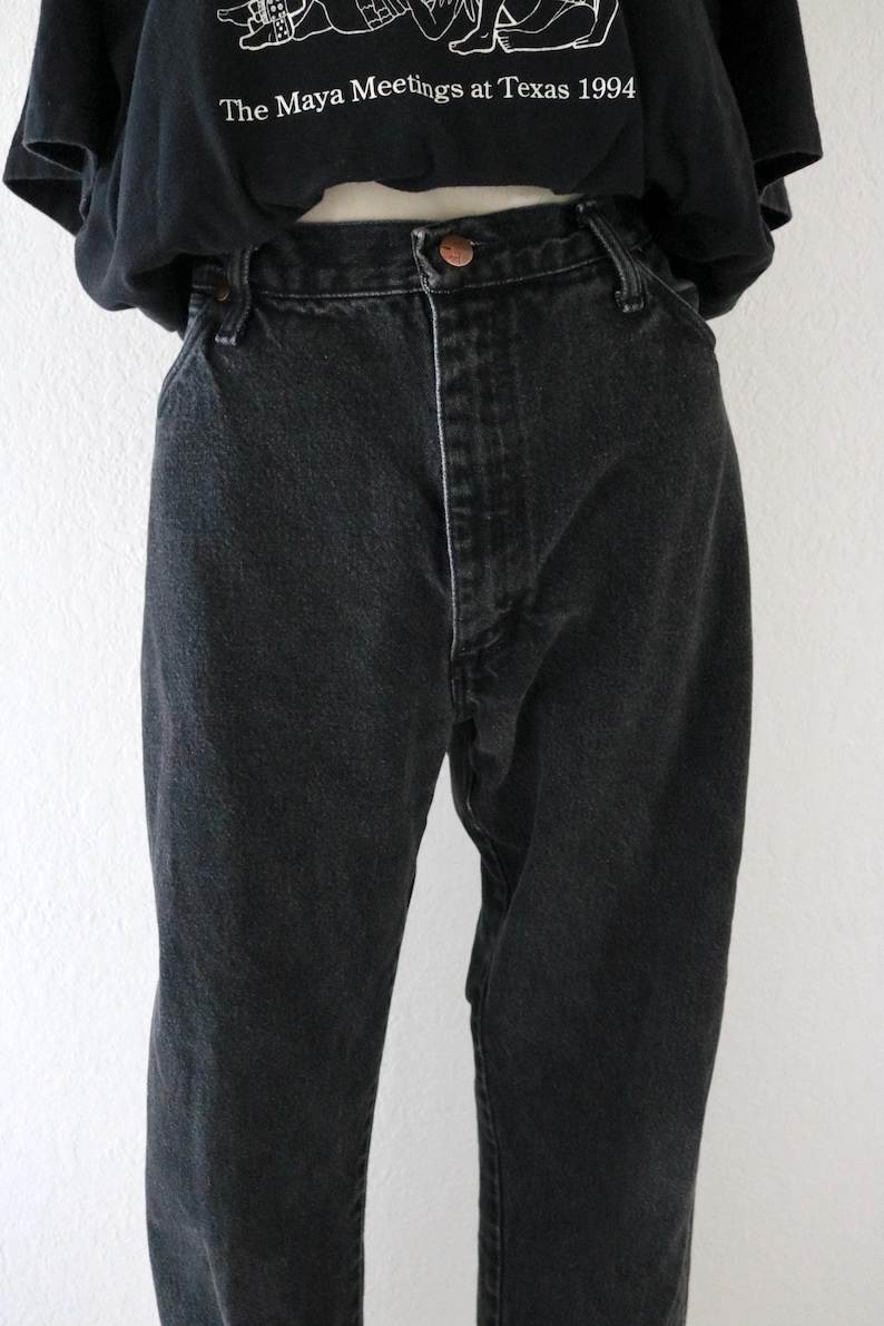 May include: Black denim jeans with a button closure and a straight leg fit. The jeans are worn with a black t-shirt that has the text "The Maya Meetings at Texas 1994" printed on it.