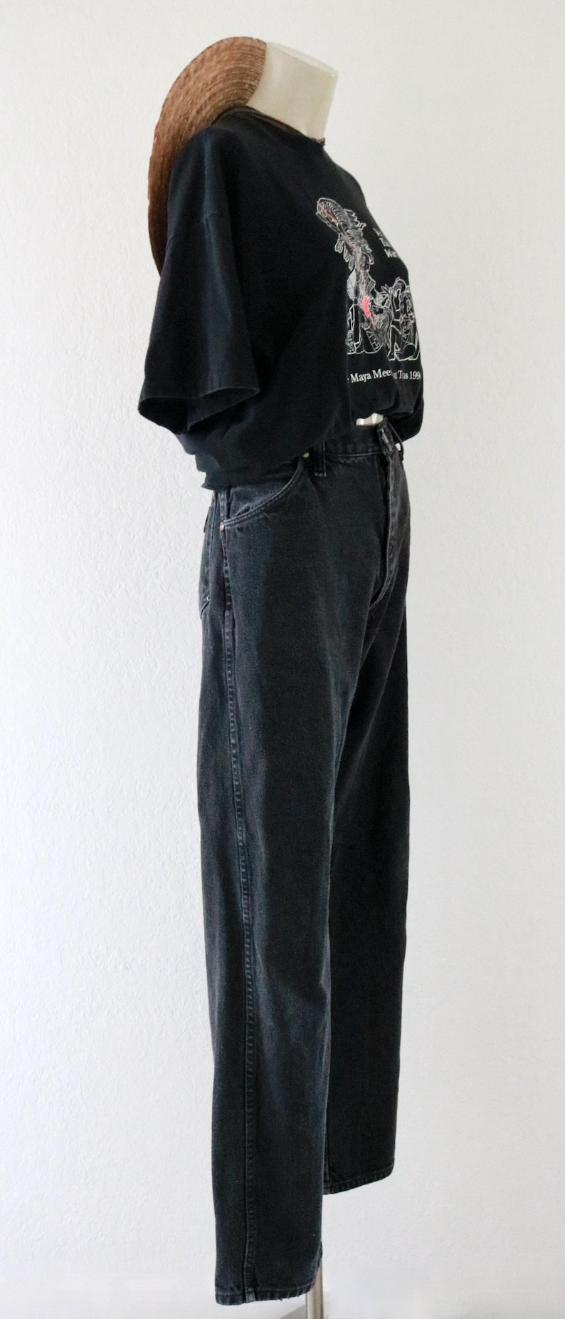 May include: A black t-shirt with a graphic design and the text "Maya Meet 199". The shirt is paired with black denim jeans and a straw hat. The outfit is displayed on a mannequin against a white background.