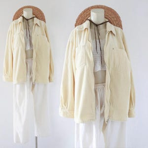 May include: A cream-colored corduroy jacket with a zipper closure, paired with white pants. The jacket has two chest pockets and is displayed on a mannequin. A straw hat is placed on top of the mannequin's head.