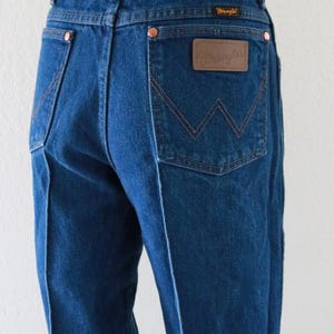 May include: Dark blue denim jeans with a high waist and a classic Wrangler logo patch on the back pocket. The jeans feature copper rivets and a stitched W design. The jeans are a vintage style.