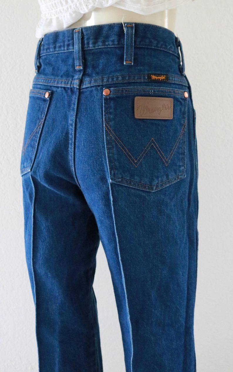 May include: Dark blue denim jeans with a high waist and a classic Wrangler logo patch on the back pocket. The jeans feature copper rivets and a stitched W design. The jeans are a vintage style.