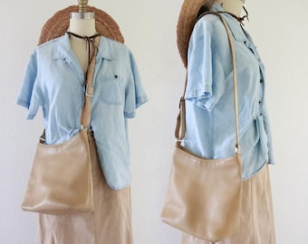 May include: A tan leather crossbody bag with an adjustable strap. The bag is displayed on a mannequin wearing a light blue denim shirt, a tan skirt, and a straw hat. The bag has a simple, classic design.