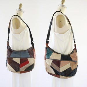 May include: A multi-colored leather hobo bag featuring a patchwork design of various geometric shapes in shades of brown, cream, black, blue, and red. The bag includes a long adjustable shoulder strap, a top zipper closure, and a front exterior pocket.