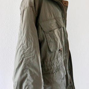 Pu&ograve; includere: Giacca verde oliva con multiple tasche frontali, colletto alto e chiusura con cerniera. La giacca ha una vestibilit&agrave; ampia e sembra essere realizzata in un tessuto leggero. La giacca &egrave; esposta su un manichino.