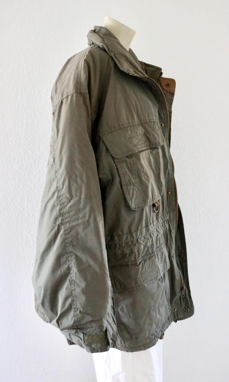Pu&ograve; includere: Giacca verde oliva con multiple tasche frontali, colletto alto e chiusura con cerniera. La giacca ha una vestibilit&agrave; ampia e sembra essere realizzata in un tessuto leggero. La giacca &egrave; esposta su un manichino.