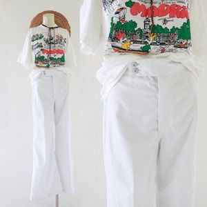 Puede incluir: Conjunto blanco con una camiseta de manga corta con un estampado gráfico colorido y pantalones blancos de pernera ancha. La camiseta tiene cuello redondo y un lazo decorativo. Los pantalones tienen cintura alta y cierre de botón. Un sombrero de paja está encima.