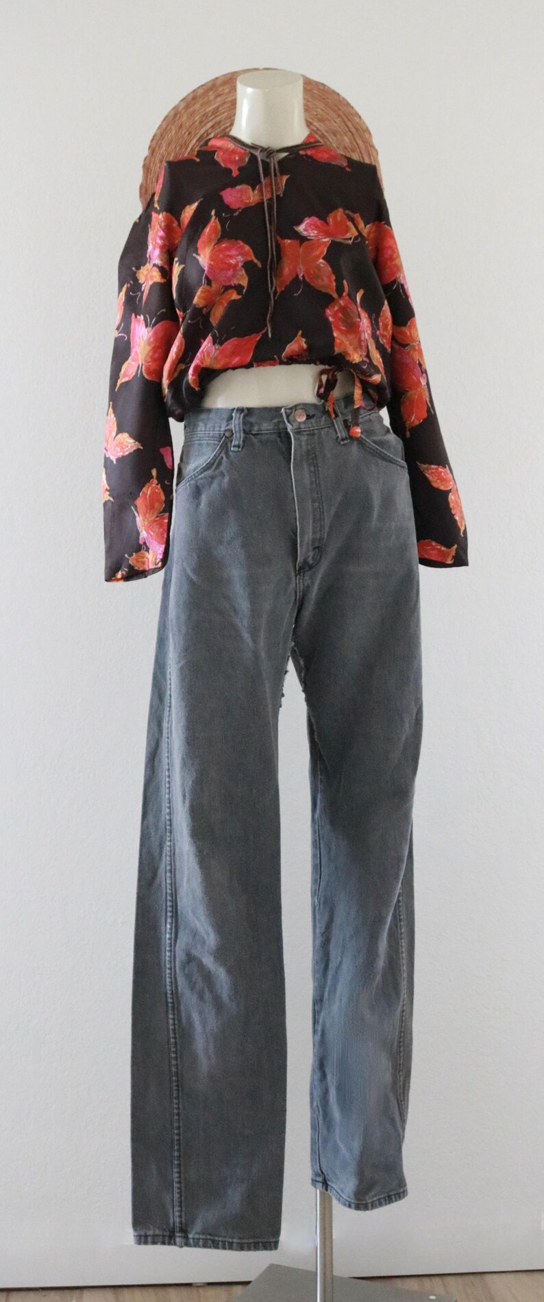 Puede incluir: Un maniqu&iacute; exhibe una blusa negra con un estampado floral rojo y naranja, combinada con vaqueros grises. Los vaqueros tienen cintura alta y pernera ancha. Un sombrero de paja est&aacute; encima.