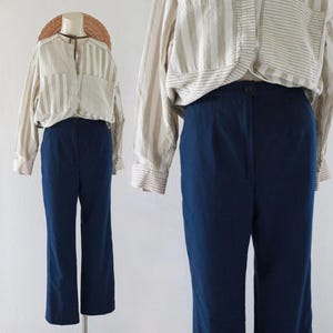 May include: A mannequin displays a navy blue pair of trousers and a striped button-up shirt. The trousers are high-waisted with a straight leg. The shirt is beige and white striped with long sleeves. The outfit is a vintage style.