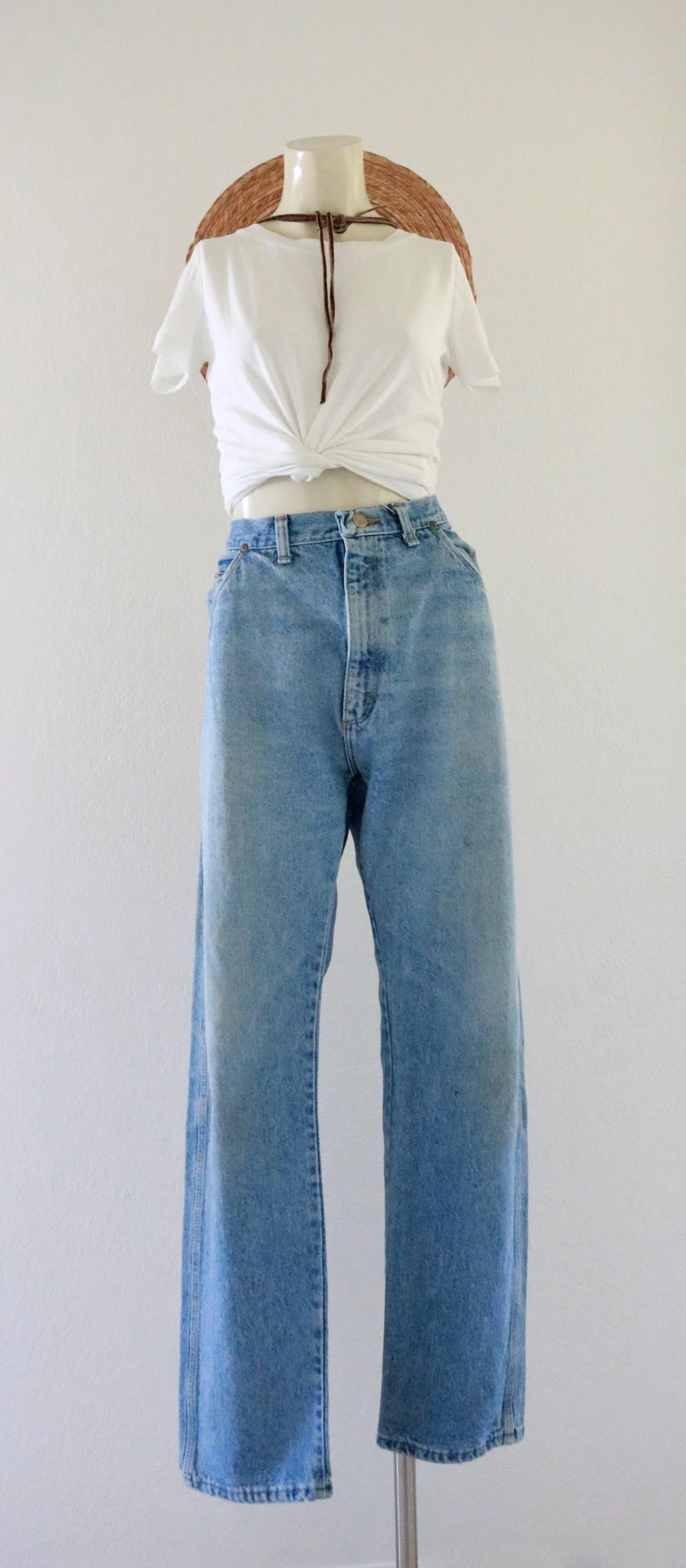 May include: A mannequin displays a casual outfit. It features light-wash denim jeans with a high waist and a white, knotted t-shirt. A straw hat with a brown cord is also included.