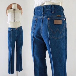 May include: Dark blue denim Wrangler jeans. The jeans have a high waist and a straight leg. The back pocket features a brown leather patch with the Wrangler logo. The jeans are displayed on a mannequin.