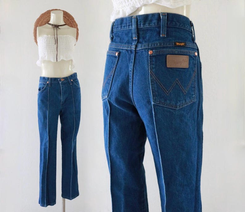 May include: Dark blue denim Wrangler jeans. The jeans have a high waist and a straight leg. The back pocket features a brown leather patch with the Wrangler logo. The jeans are displayed on a mannequin.