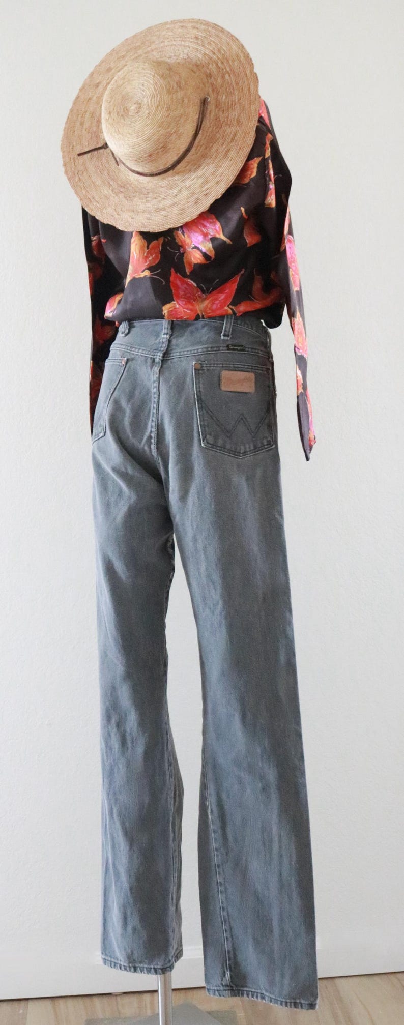 Puede incluir: Una persona con un sombrero de paja y una blusa negra con un estampado floral rojo. La persona lleva vaqueros Wrangler de mezclilla gris.