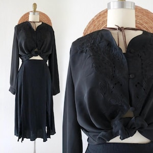 以下が含まれることがあります： 黒色のツーピースの衣装。長袖ブラウスは、複雑な花の刺繍と結び目のフロントが特徴です。お揃いのスカートはミディ丈でスリット入り。麦わら帽子をかぶったマネキンに飾られています。