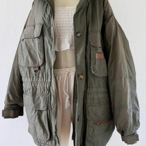 Pu&ograve; includere: Giacca verde oliva con pi&ugrave; tasche, chiusura con cerniera e dettagli con bottoni. La giacca ha un punto vita con coulisse e un collo alto. La giacca &egrave; esposta su un manichino con top e pantaloni bianchi.