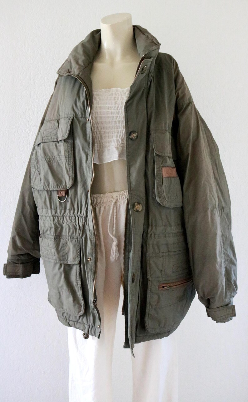Pu&ograve; includere: Giacca verde oliva con pi&ugrave; tasche, chiusura con cerniera e dettagli con bottoni. La giacca ha un punto vita con coulisse e un collo alto. La giacca &egrave; esposta su un manichino con top e pantaloni bianchi.