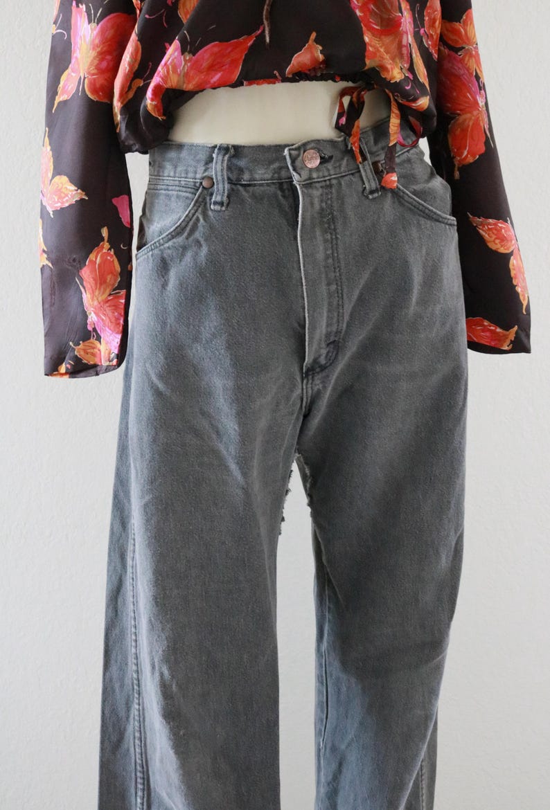 Puede incluir: Vaqueros grises de mezclilla con cintura alta y pernera recta. Los vaqueros tienen el estilo cl&aacute;sico de cinco bolsillos y cierre de bot&oacute;n y cremallera. La modelo lleva una blusa negra con estampado floral.
