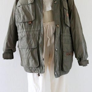 Pu&ograve; includere: Giacca verde oliva con pi&ugrave; tasche, vita con coulisse e chiusura con bottoni. La giacca &egrave; abbinata a pantaloni bianchi e un top bianco. La giacca ha un cappuccio e maniche lunghe.