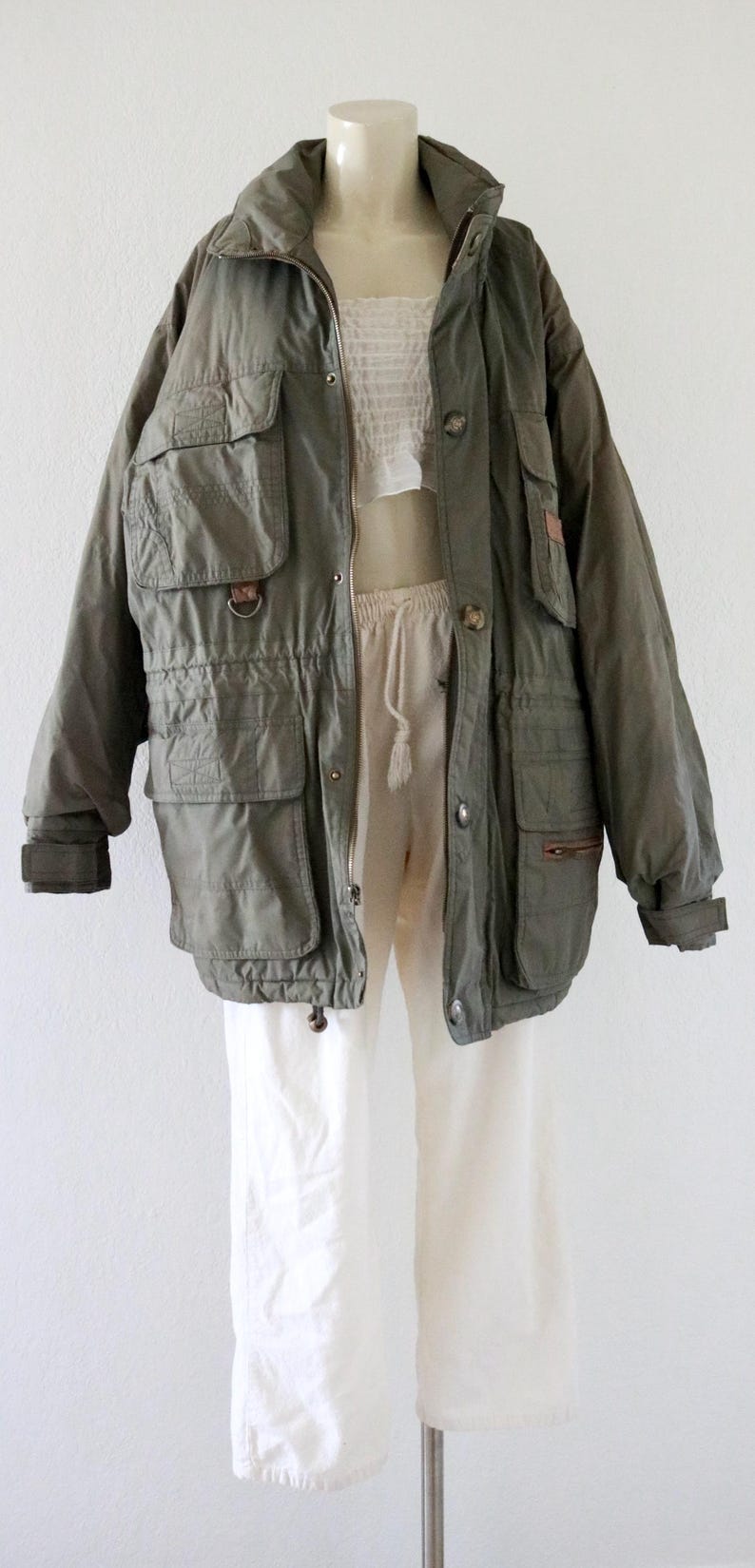 Pu&ograve; includere: Giacca verde oliva con pi&ugrave; tasche, vita con coulisse e chiusura con bottoni. La giacca &egrave; abbinata a pantaloni bianchi e un top bianco. La giacca ha un cappuccio e maniche lunghe.