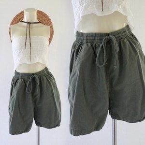 May include: Olive green shorts featuring an elastic waistband with a matching drawstring tie. The shorts have a relaxed fit and side pockets. They are displayed on a mannequin alongside a white strapless tube top and a straw hat.