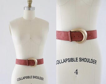 May include: A dusty rose-colored belt with a large, gold-toned buckle. The belt is made of a suede-like material and is displayed on a mannequin. The buckle is a half-moon shape. The belt is a fashion accessory.