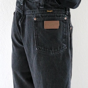 May include: Black denim Wrangler jeans. The jeans have a classic five-pocket design, belt loops, and a brown leather patch with the Wrangler logo on the back pocket. The jeans are a dark wash and appear to be vintage.