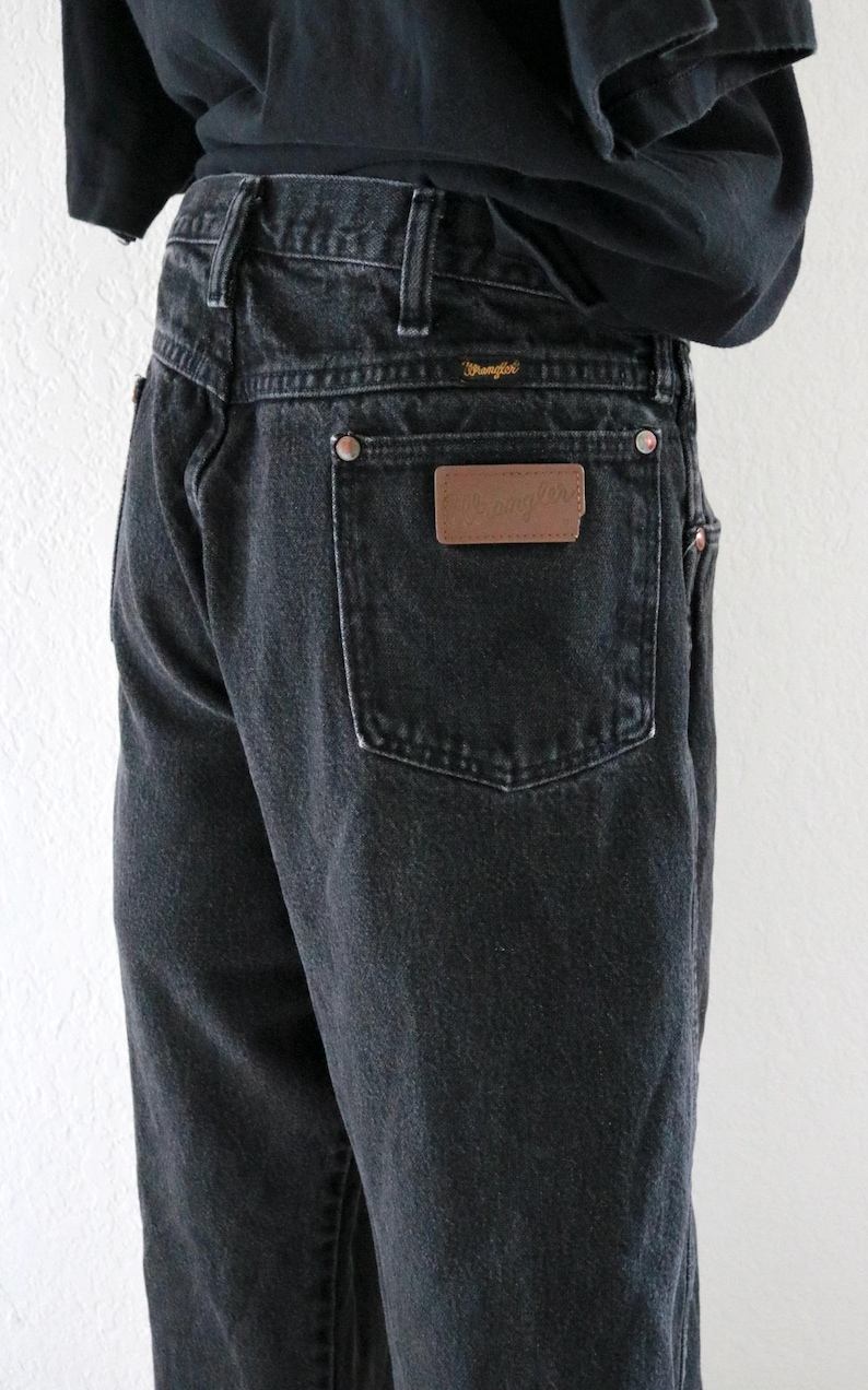 May include: Black denim Wrangler jeans. The jeans have a classic five-pocket design, belt loops, and a brown leather patch with the Wrangler logo on the back pocket. The jeans are a dark wash and appear to be vintage.